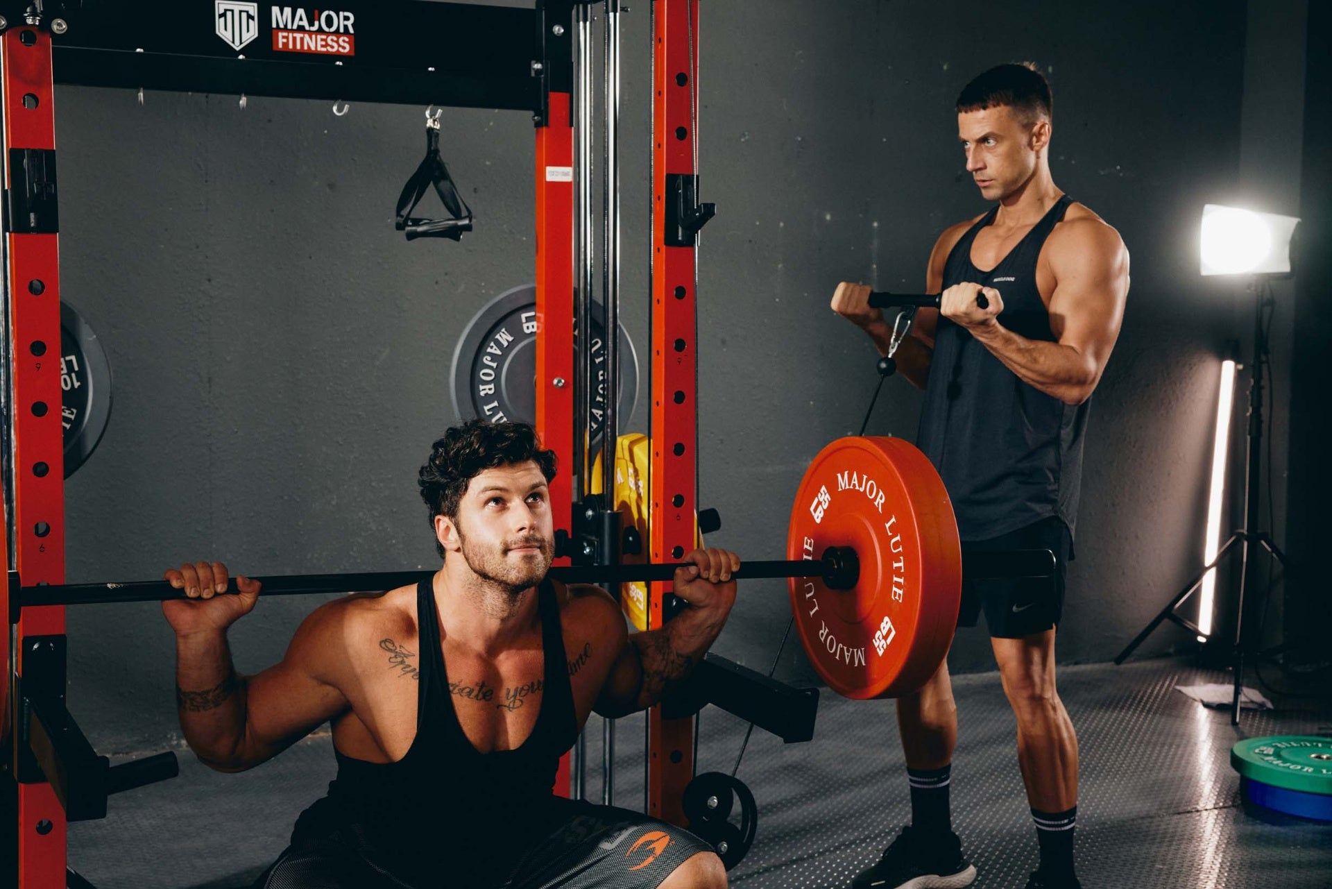 Two men using a power rack in a home gym, one squatting and the other doing a cable curl.
